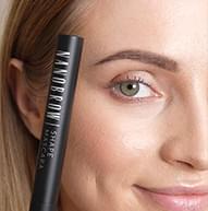 Modelka uśmiecha się, trzymając tusz do rzęs Nanobrow Shape blisko ucha