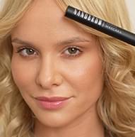 Modelka blondynka, uśmiechnięta, trzymająca tusz do rzęs Nanobrow Shape Mascara blisko czoła